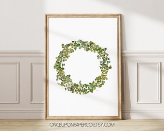 May include: A framed print featuring a watercolor wreath composed of green and yellow-green leaves. The wreath is centered on a white background, and the frame is a light brown color. The Etsy shop name is visible at the bottom.