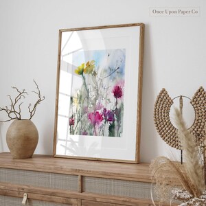 May include: Watercolor painting of a floral bouquet with yellow, pink, and purple flowers. The painting is framed in a natural wood frame and is hanging on a wall.