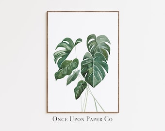 May include: Watercolor painting of a Monstera plant with large, green, split leaves. The artwork is framed in a brown wooden frame and set against a white background. The text "Once Upon Paper Co" is printed at the bottom.