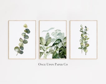 May include: Set of three watercolor prints featuring green foliage, including eucalyptus and monstera leaves. The prints are framed in a simple wooden frame and have a white background. The text "Once Upon Paper Co" is printed below the prints.