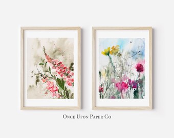 May include: Two framed watercolor paintings of flowers. One features a tall, pink flowering plant with green leaves against a muted background. The other displays a variety of colorful flowers, including yellow and pink, with a blue sky backdrop. The text "Once Upon Paper Co" is at the bottom.