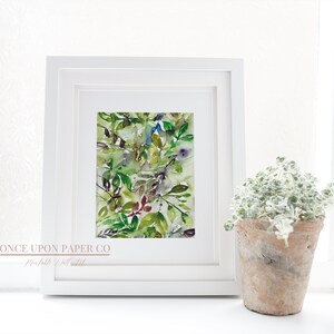 May include: A framed watercolor painting of green, blue, and purple leaves. The painting is in a white frame and is sitting on a white surface. The text "ONCE UPON PAPER CO" is visible at the bottom of the frame.