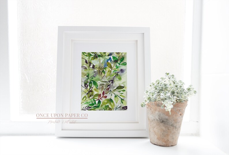 May include: A framed watercolor painting of green, blue, and purple leaves. The painting is in a white frame and is sitting on a white surface. The text "ONCE UPON PAPER CO" is visible at the bottom of the frame.