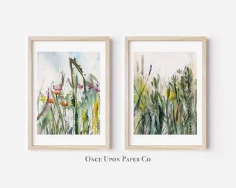 May include: Two framed watercolor paintings of wildflowers. The artwork features a variety of colorful flowers and grasses in shades of green, yellow, orange, and purple. The frames are light-colored wood.