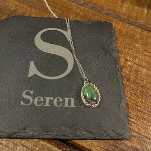 May include: A silver chain necklace with a green and pink oval gemstone pendant. The pendant is set in a silver bezel with a decorative edge. The necklace is on a black slate surface with the word "Seren" engraved on it.