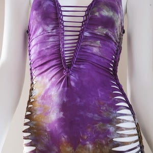 Peut inclure: Un body sans manches tie-dye dans les tons de violet, marron et blanc. L'encolure en V et les côtés présentent des détails découpés et tressés. Le vêtement est présenté sur un mannequin.