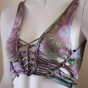 May include: A tie-dye crop top in shades of purple, green, and blue. The top features a V-neck and braided detailing across the chest and straps. The fabric has a soft, flowing appearance.
