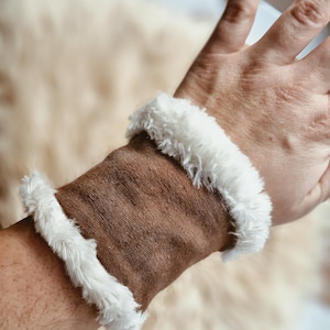 Op de afbeelding: Bruine polsband met witte imitatiebont afwerking. De manchet is gemaakt van een textuurstof en is ontworpen om om de pols te worden gedragen. De witte bontafwerking geeft het accessoire een zacht, gezellig uiterlijk.