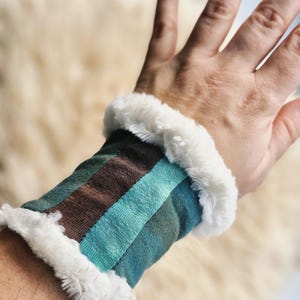 Op de afbeelding: Een polsband met een gestreept patroon in bruin, blauwgroen en blauw, afgezet met pluizig wit imitatiebont. De polsband is om een pols gewikkeld en toont het ontwerp en de textuur van de stof.