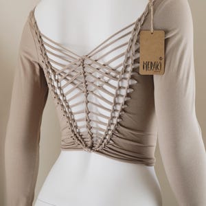 May include: Tan long-sleeve top with a unique open back design. The back features a woven lattice pattern. A brown tag with the word "MERAKI" is attached to the shoulder. The top is displayed on a white mannequin.