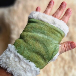 Può includere: Un guanto senza dita verde e bianco. Il corpo principale del guanto è in tessuto verde con un motivo sottile e i bordi sono rifiniti con soffice ecopelliccia bianca. Il guanto è progettato per coprire la mano.