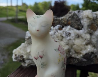 May include: A ceramic cat figurine with a cream-colored body and pink accents. The cat is decorated with hand-painted pink and yellow flowers and green leaves. The cat is sitting on a wooden surface.