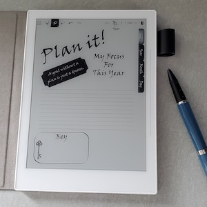 May include: A white e-reader with a black and white handwritten note that says "Plan it!" and "A goal without a plan is just a dream." The note also says "My Focus For This Year." There is a blank space labelled "Key" with a small key icon. The e-reader has a side bar with the words "Home", "Year", "Month", and "Day".