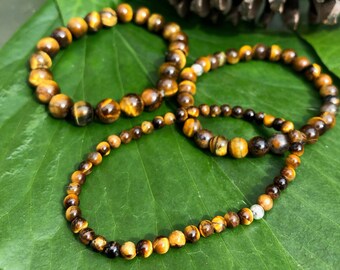 May include: Three brown tiger's eye beaded bracelets on a green leaf background. The bracelets are made of small, round beads and have a silver bead accent.