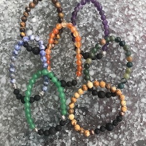 Può includere: Sette braccialetti di perline con pietre di diversi colori e perline di lava. I braccialetti sono disposti in cerchio su uno sfondo grigio.