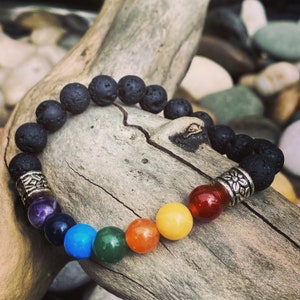 Puede incluir: Un brazalete de roca de lava negra con detalles plateados y siete cuentas de colores de chakra. El brazalete está sobre un trozo de madera a la deriva.