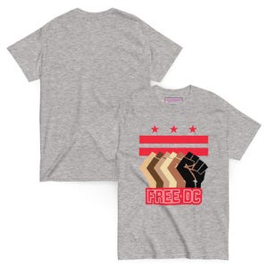 Könnte beinhalten: Graues T-Shirt mit einem grafischen Design. Das Design umfasst rote Streifen, drei rote Sterne und eine Reihe stilisierter Fäuste in verschiedenen Hauttönen. Der Text "FREE DC" befindet sich in einem roten Feld.