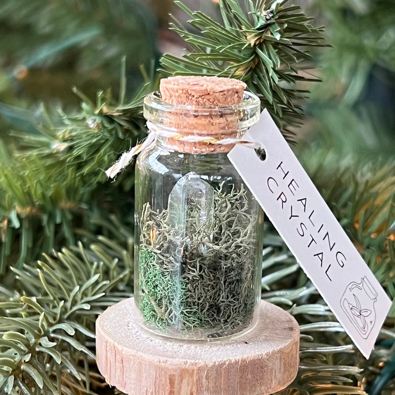 May include: A small glass jar with a cork lid filled with green moss and a clear quartz crystal. A white tag is attached to the jar with the text "HEALING CRYSTAL".