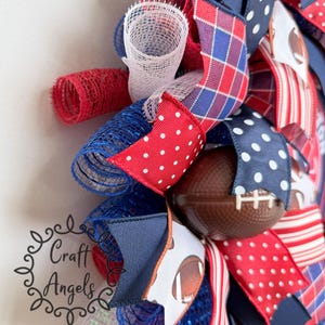 May include: A decorative wreath featuring red, white, and blue mesh loops and ribbons with polka dot, plaid, and striped patterns. The wreath includes miniature brown footballs with white laces and a logo reading Craft Angels in black script.