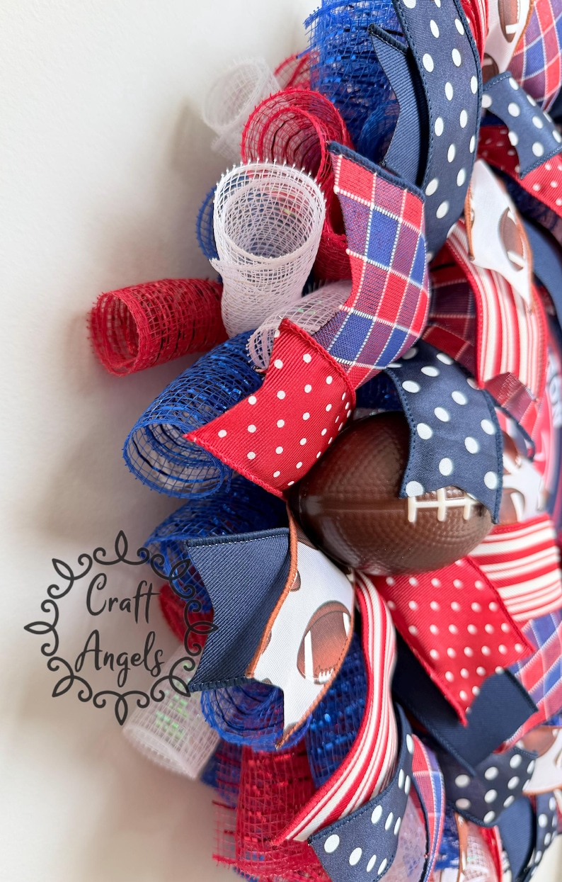 May include: A decorative wreath featuring red, white, and blue mesh loops and ribbons with polka dot, plaid, and striped patterns. The wreath includes miniature brown footballs with white laces and a logo reading Craft Angels in black script.
