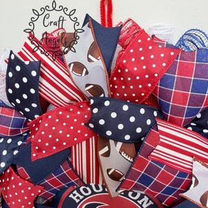 May include: A decorative wreath featuring red, white, and blue ribbons with polka dot, striped, and plaid patterns. Some ribbons display a football graphic. A circular sign with the text HOUSTON is visible at the bottom, and a red pipe cleaner hanger.