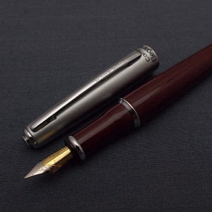 Op de afbeelding: Een zilveren en bordeauxrode vulpen met de woorden "KOKUYO CAMIO Elegant" gegraveerd op de dop.