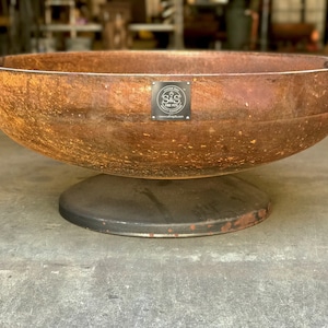 May include: A large, rusty metal fire pit with a black metal base. The fire pit has a round bowl shape and a raised lip. A black metal tag is attached to the side of the bowl with the text "Custom Built Fire Pit" and a logo. The text "www.sdfirepits.com" is also on the tag.