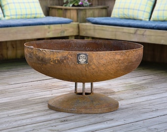 May include: A large, rusty metal fire pit bowl with a round base and three legs. The bowl is sitting on a wooden deck.