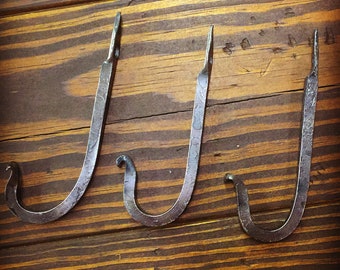 May include: Three rustic metal hooks, each with a unique curve, are mounted on a wooden surface. The hooks are made of dark metal and have a hammered finish.