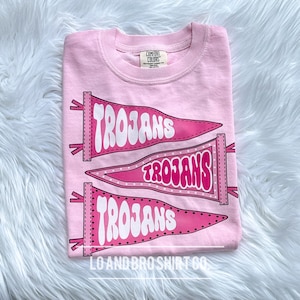 May include: Pink t-shirt featuring three pink pennants with the word "Trojans" in white. The shirt has a small tag that reads "Comfort Colors" and the bottom of the shirt says "Lo and Bro Shirt Co."