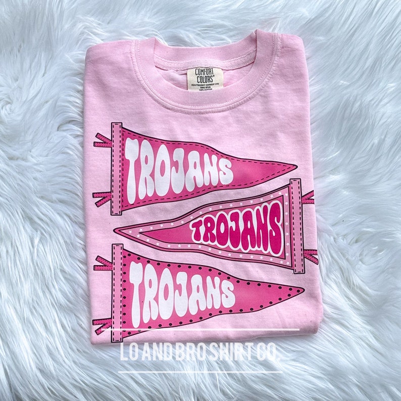 May include: Pink t-shirt featuring three pink pennants with the word "Trojans" in white. The shirt has a small tag that reads "Comfort Colors" and the bottom of the shirt says "Lo and Bro Shirt Co."