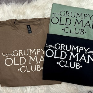 May include: Three t-shirts in brown, sage green, and black, each with the text "GRUMPY OLD MAN CLUB" in white. The text is in a stylized font with decorative flourishes. The shirts are folded and arranged on a fluffy white surface.