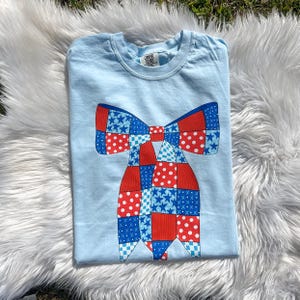 Puede incluir: Una camiseta azul claro con un diseño de corbata de lazo de patchwork rojo, blanco y azul. La corbata de lazo está hecha de cuadrados con diferentes patrones, incluyendo estrellas, lunares y cuadros. La camiseta tiene cuello redondo y mangas cortas.
