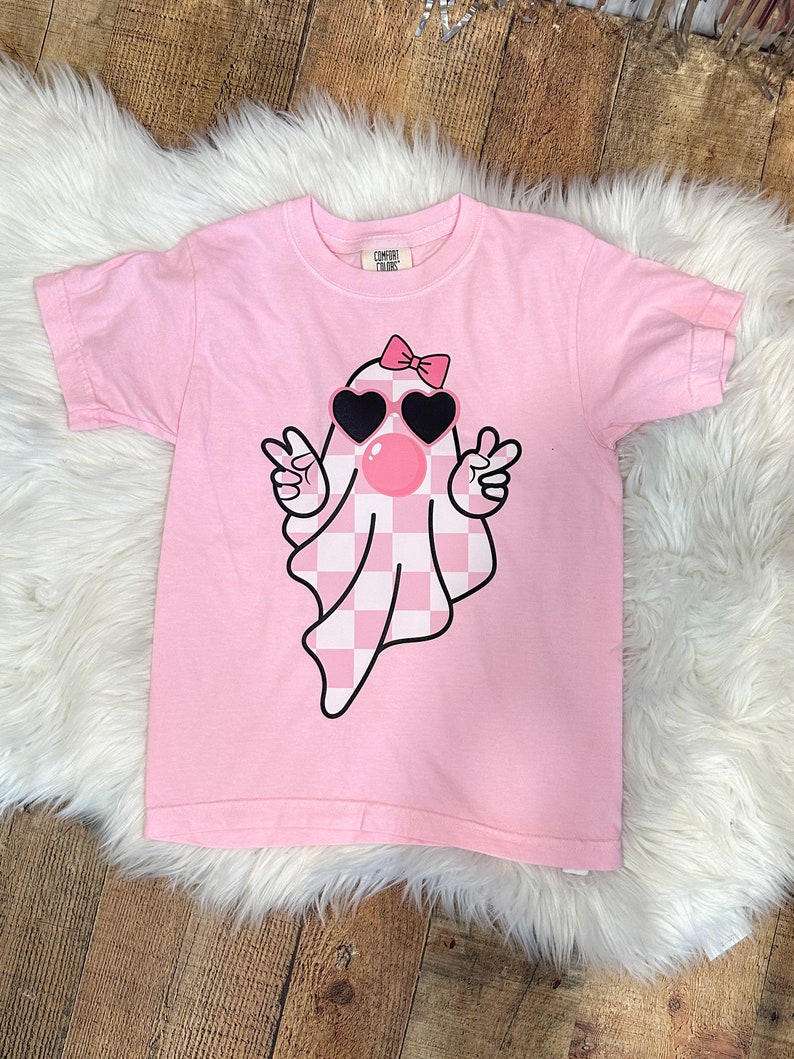以下が含まれることがあります： ピンクの半袖Tシャツに、サングラスとリボンをつけた幽霊のグラフィックがプリントされています。幽霊はピンクと白のチェック柄です。