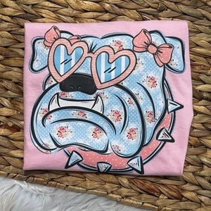 May include: Pink t-shirt with a cartoon bulldog design. The bulldog has heart-shaped sunglasses and a pink bow. The design features a blue and white floral pattern with pink accents. The shirt is on a woven background.