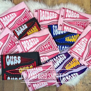 May include: A collection of pink and purple t-shirts with various sports team names printed on them. The names include "Bobcats", "Bears", "Pirates", "Cougars", "Cubs", "Trojans", "Raiders", "Falcons", "Knights", "Toros", "Wildcats", and "Bobcats".
