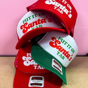 Könnte beinhalten: Ein Stapel roter, weißer und grüner Trucker-Hüte mit dem Text "PUT IT ON Santa's TAB" in einem Retro-Schriftstil. Die Hüte haben einen Mesh-Rücken und eine Schaumstofffront. Die Hüte sind übereinander gestapelt.