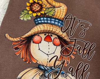 May include: Brown t-shirt with a cartoon scarecrow wearing a straw hat with a sunflower on top. The text "It's Fall Y'all" is written in white cursive.