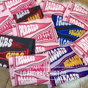 May include: A collection of pink, purple and grey t-shirts with various school team names printed on them. The names include "Cubs", "Trojans", "Pirates", "Wildcats", "Falcons", "Knights", "Bears", "Cougar", "Bobcats", "Raiders", and "Toros".