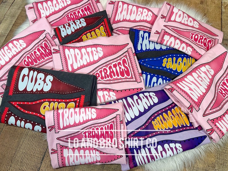 May include: A collection of pink, purple and grey t-shirts with various school team names printed on them. The names include "Cubs", "Trojans", "Pirates", "Wildcats", "Falcons", "Knights", "Bears", "Cougar", "Bobcats", "Raiders", and "Toros".