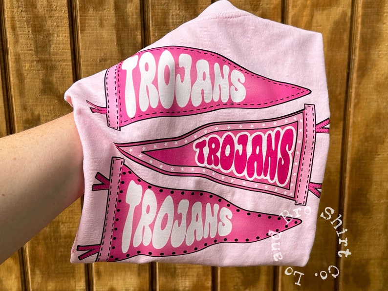 May include: Pink t-shirt featuring three pink pennants with the word "Trojans" in white lettering. The shirt has a black outline around the pennants and text. The shirt is folded and held by a hand.