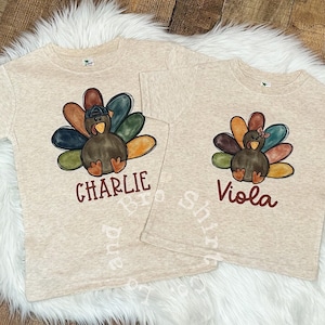 Può includere: Due magliette beige con un design di tacchino del Ringraziamento. La maglietta a sinistra ha il nome "Charlie" stampato sotto il tacchino. La maglietta a destra ha il nome "Viola" stampato sotto il tacchino.