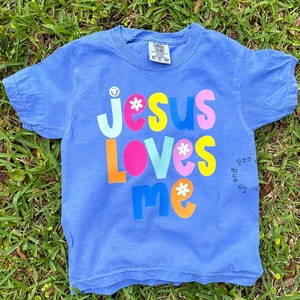 May include: A blue t-shirt featuring the phrase "Jesus Loves Me" in bright, multi-coloured letters with flower and smiley face details. The shirt is displayed on a bed of green grass.