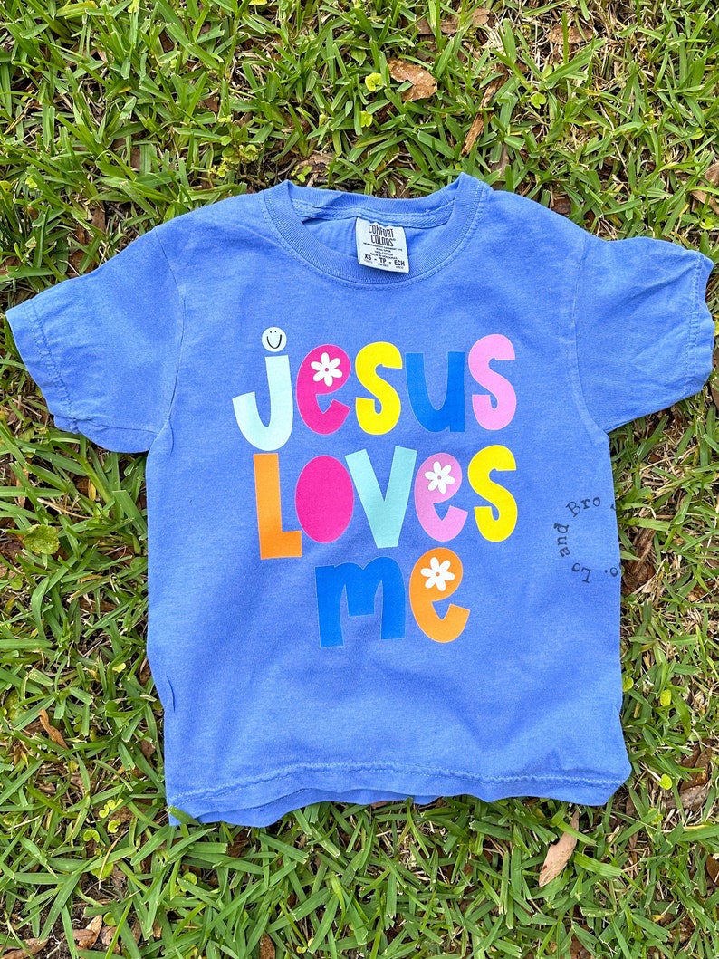 May include: A blue t-shirt featuring the phrase "Jesus Loves Me" in bright, multi-coloured letters with flower and smiley face details. The shirt is displayed on a bed of green grass.