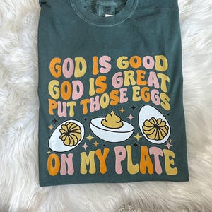 May include: Green t-shirt with the text "God is good God is great Put those eggs on my plate" in a retro font. The text is surrounded by three cartoon images of deviled eggs.