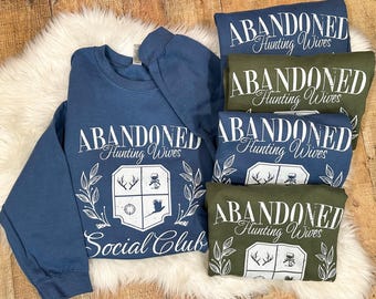 Puede incluir: Cuatro sudaderas azules y verdes con el texto "Abandoned Hunting Wives Social Club" y un escudo con un ciervo, una brújula, un pájaro y una copa de vino.