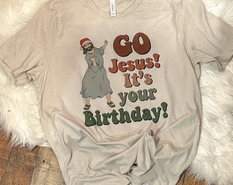 Op de afbeelding: Een beige T-shirt met een cartoon afbeelding van Jezus die een kerstmanmuts draagt en danst. De tekst "GO Jesus! It's your Birthday!" staat op het shirt gedrukt.