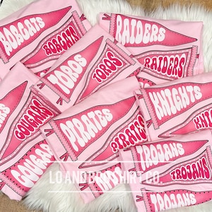 May include: Pink t-shirts featuring a range of school team pennant designs. The designs include "Bobcats", "Cougar", "Toros", "Pirates", "Raiders", "Knights", and "Trojans".