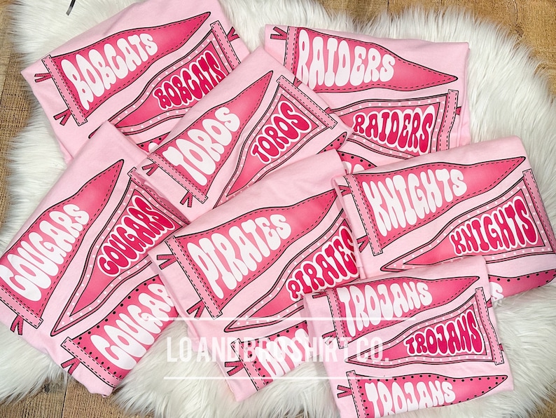 May include: Pink t-shirts featuring a range of school team pennant designs. The designs include "Bobcats", "Cougar", "Toros", "Pirates", "Raiders", "Knights", and "Trojans".