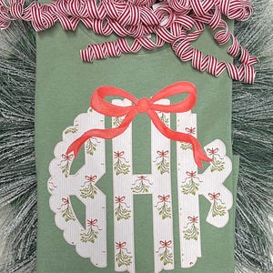 May include: A green t-shirt featuring a white and red monogram design. The monogram is encircled by a wreath of mistletoe and bows. The shirt is adorned with a red bow.